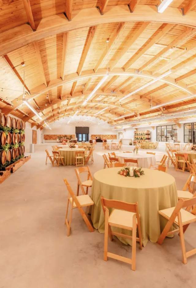 Interior of the Muellner Building event space, featuring a vaulted wood-beam ceiling, rows of decorative wine barrels, string lights and round banquet tables set for an event.