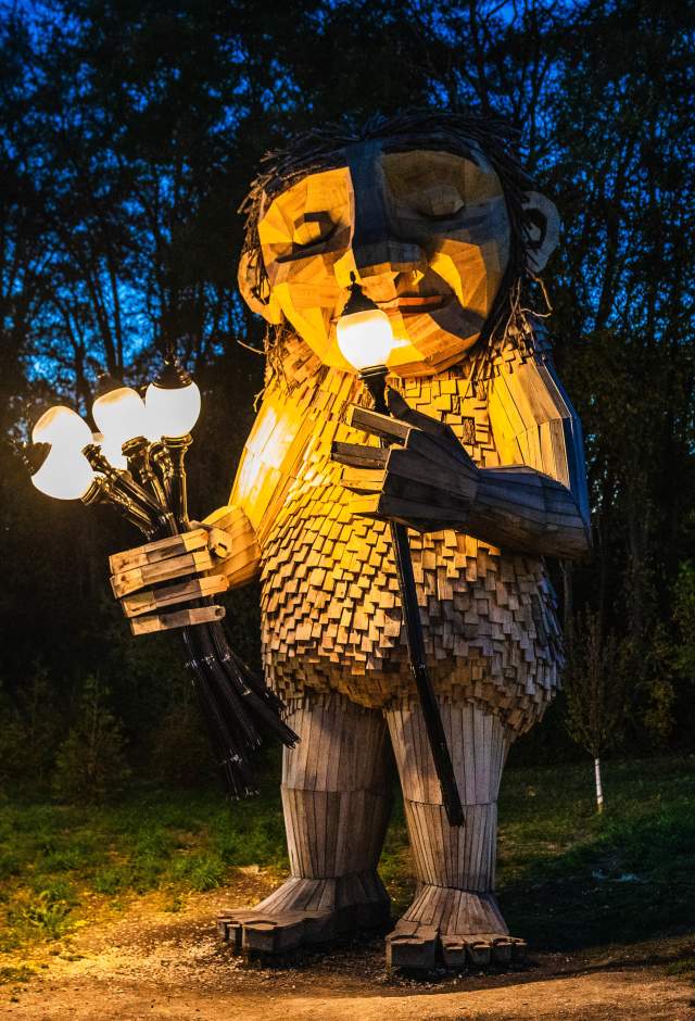 The Thomas Dambo Troll, Mama Rosa is a large wooden sculpture which stands outdoors at dusk, illuminated by warm lights it holds in its hands. The figure is made of textured wooden planks, with a gentle, expressive face, and is surrounded by dark trees in the background.