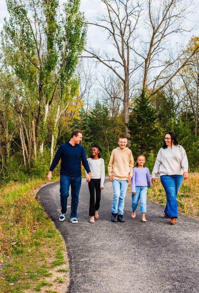 A family of five smiles and walks together along a paved trail at Firefly Grove Park surrounded by colorful autumn trees.