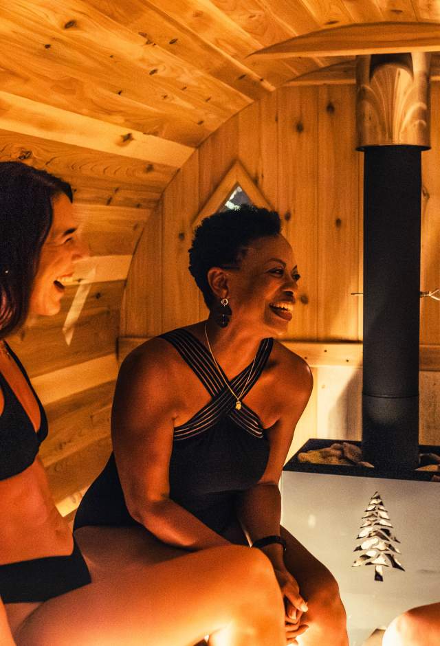 Two women in swimwear sit inside a warm, wood-paneled sauna at Heat Haven in Wauwatosa, smiling and talking near a small heater that glows softly in the center of the room.
