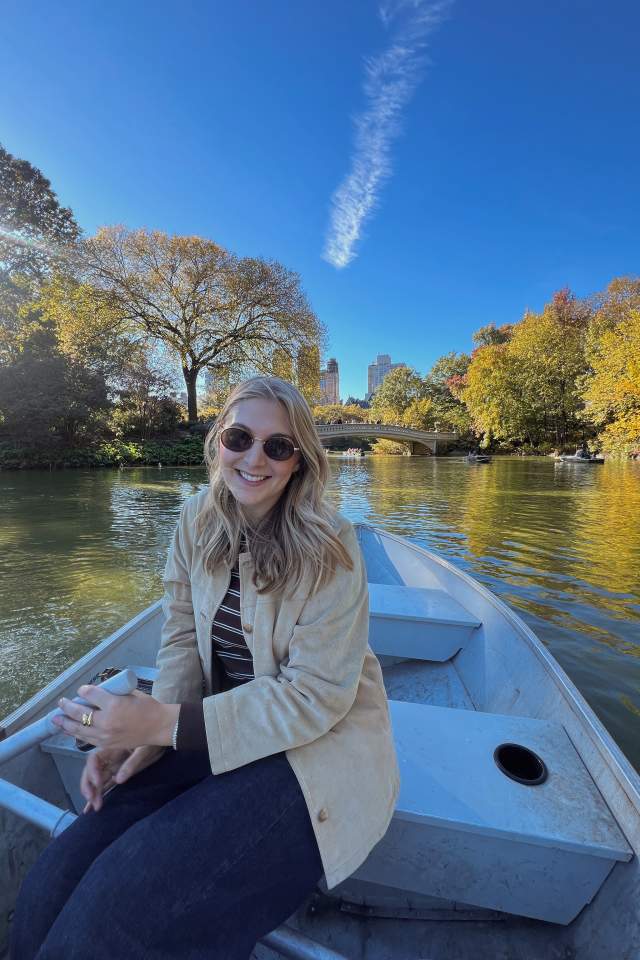 A woman in sunglasses smiles while sitting in a rowboat on a calm lake, with autumn trees and a bridge behind her under a bright blue sky.