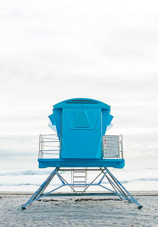 Blue lifeguard tower positioned on a sandy beach in Dania Beach, with palm trees nearby, gentle ocean waves in the background, and a long fishing pier extending into the water.