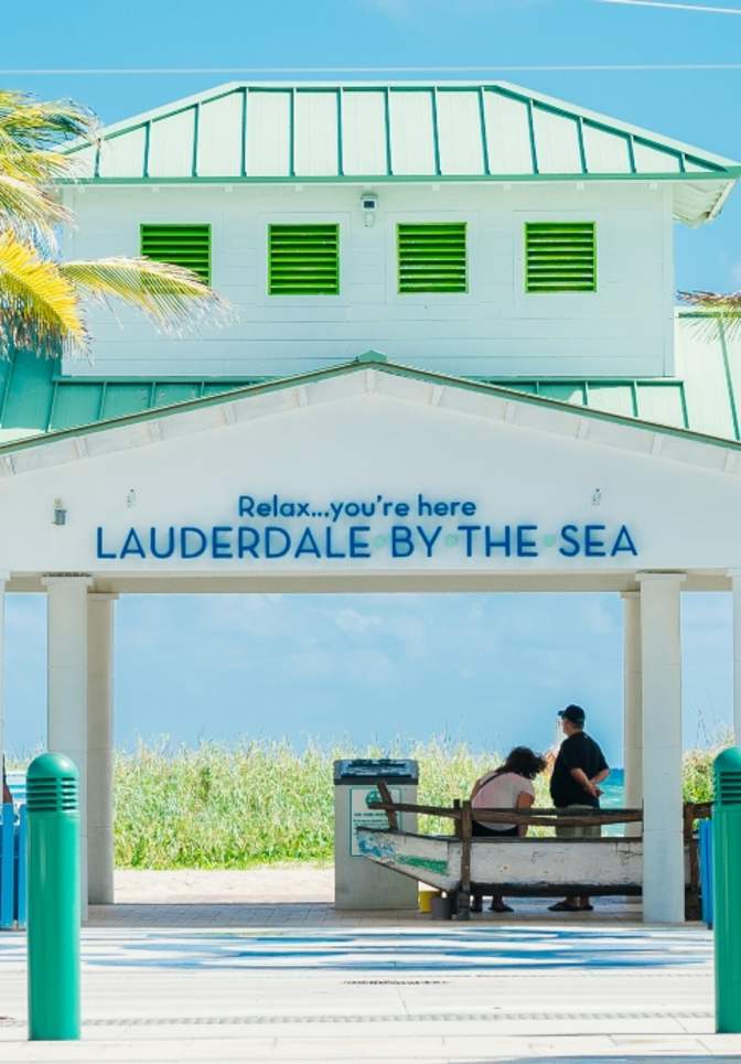 Colorful beach chairs and umbrellas line a pedestrian plaza in Lauderdale‑By‑The‑Sea, leading toward a white beachfront pavilion with palm trees and ocean views in the background.