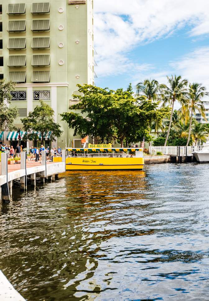 Fort Lauderdale waterfront scene with people on a dock, a yellow water taxi, palm trees, and boats docked near buildings under a partly cloudy sky.