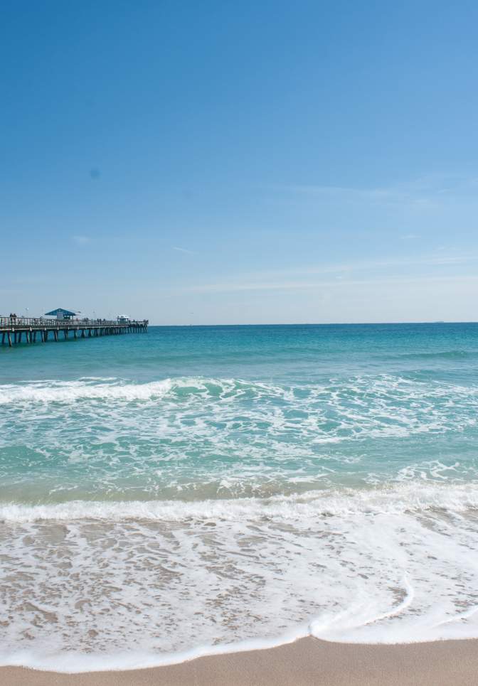 Ocean waves gently crashing onto the Lauderdale-By-The-Sea beach with a long wooden pier extending into the clear blue water under a sunny sky.