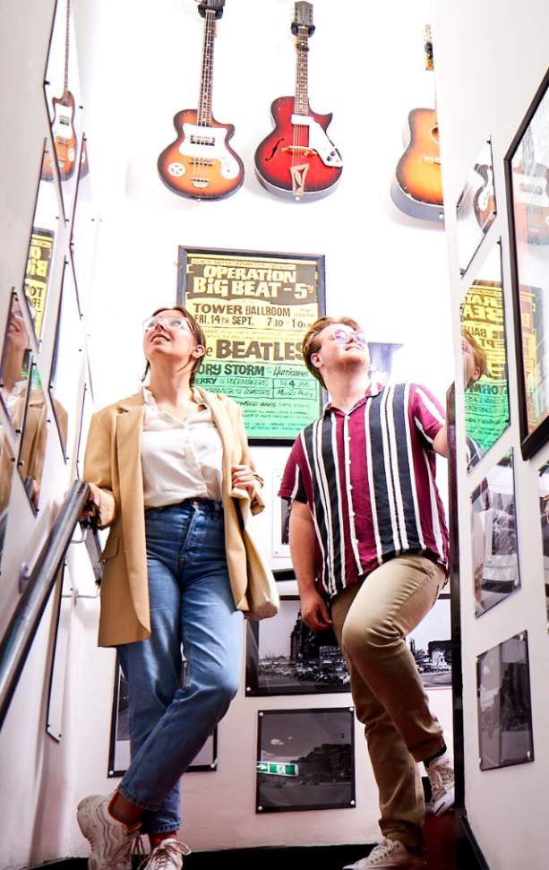 Two young people stand in the stairway of the Beatles Museum lined with Beatles posters and guitars.