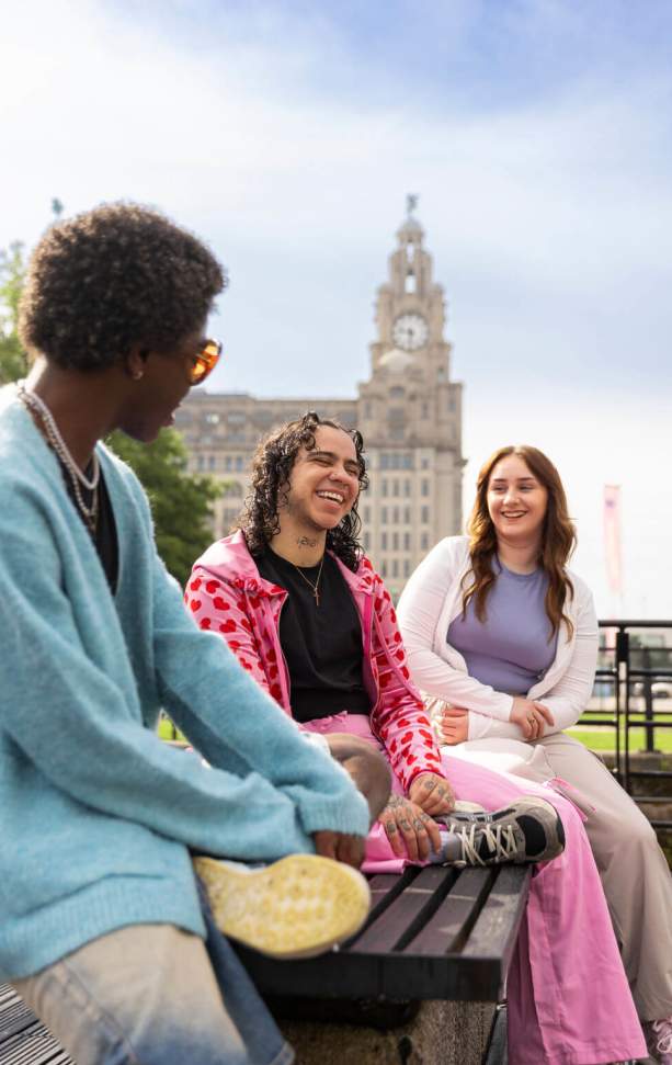 3 people sit on a bench on Liverpool Waterfront with the Royal Liver Building in the background