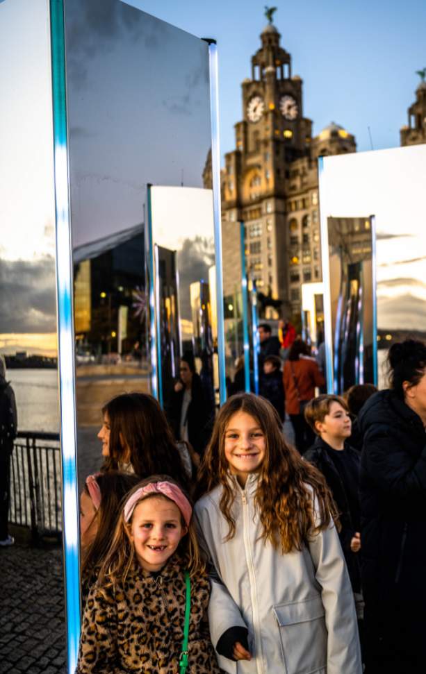 2 young girls stand amongst mirrored monoliths on Liverpool Waterfront, the Royal Liver Building can be seen in the background