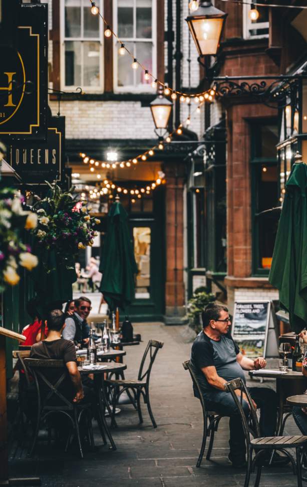 Queen avenue, a narrow Victorian Street in Liverpool City Centre. There is golden festoon lighting hung across the buildings and people outside eating at little tables and chairs.