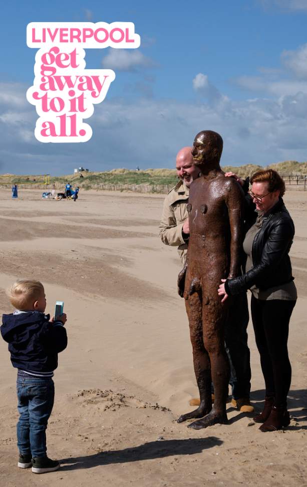 A young child is taking a picture of an adult male and female with a cast-iron, life-size statue on Crosby Beach.