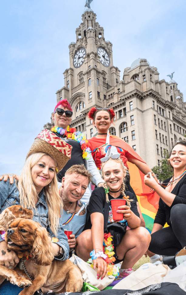 A crowd of people dressed in Pride flags and accessories on Liverpool Pier Head in front of the Liver Building