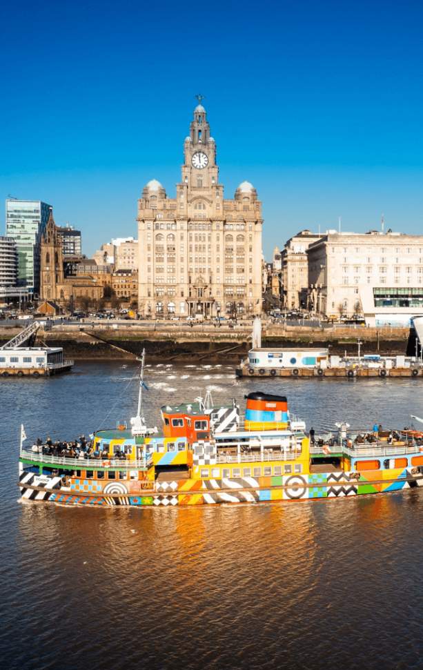 A sunny day in Liverpool. The image shows the Mersey Ferry which has been painted in bright colours and patterns on the River Mersey in front of the Royal Liver Building.