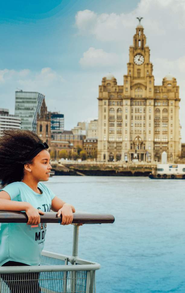 A child stands with her hands on the rail of the Mersey Ferry and overlooks the water. The Royal Liver Building can be seen in the background.