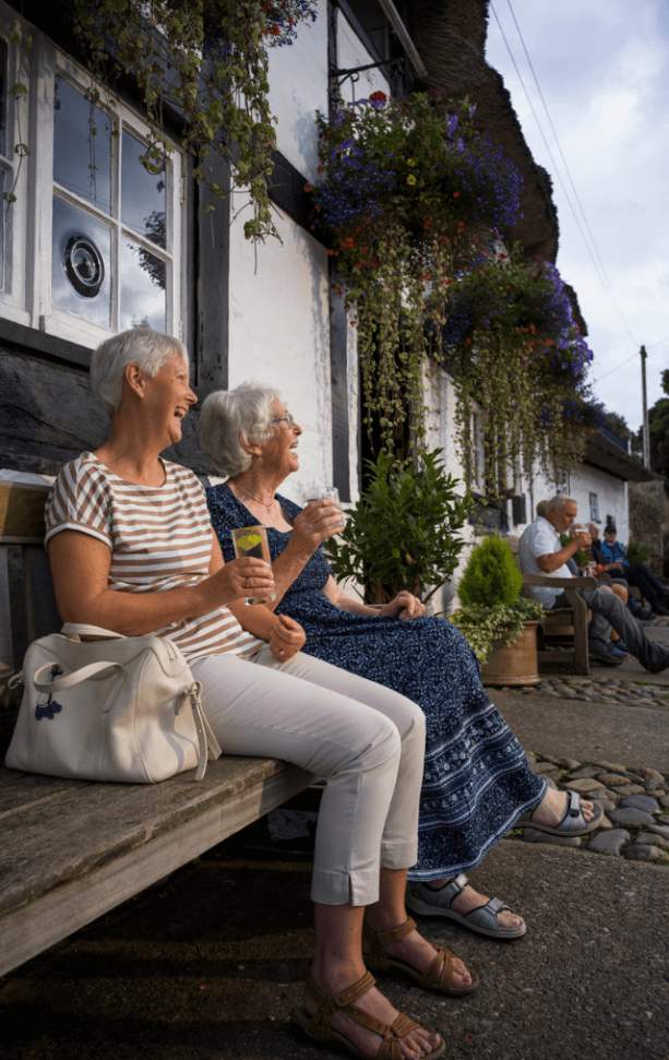 Two older women are sitting on a bench outside an old style pub drinking from glasses