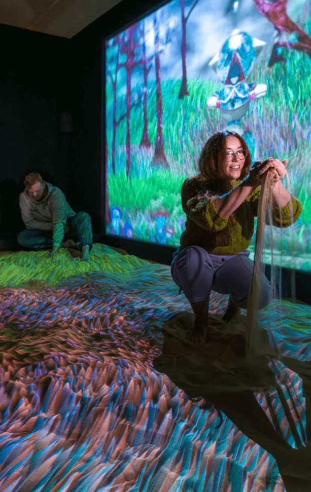 A girl picks up sand in front of a colourful projection at Subterranean Elevator