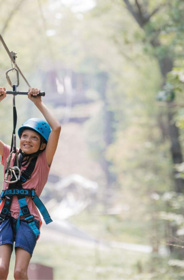 A smiling child in a blue helmet and safety harness rides a zip line through a sunlit forest, arms raised to grip the handle. The scene feels joyful and adventurous.