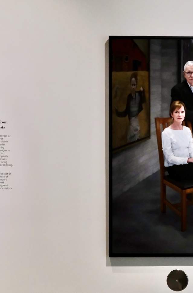 Painting displayed at Bennett Schmidt Pavilion at Muskegon Museum of Art, showing Steven Alan Bennett and Dr. Elaine Melotti Schmidt, one seated and one standing, in a formal pose against a dark backdrop. Information about the collection is presented on an adjacent wall.