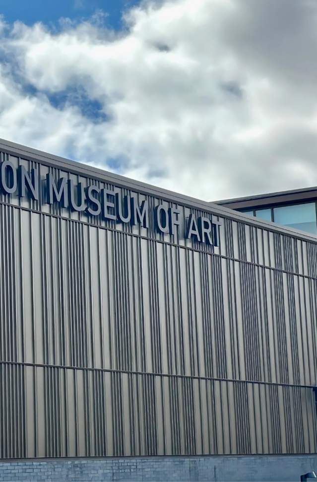 Exterior view of the Muskegon Museum of Art under a partly cloudy sky. The building features a modern design with elongated vertical panels and large glass windows.