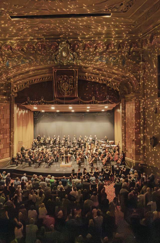 Interior of the Frauenthal Center in downtown Muskegon with a West Michigan Symphony performing on stage, warmly illuminated by golden lights with a captivated audience in attendance.