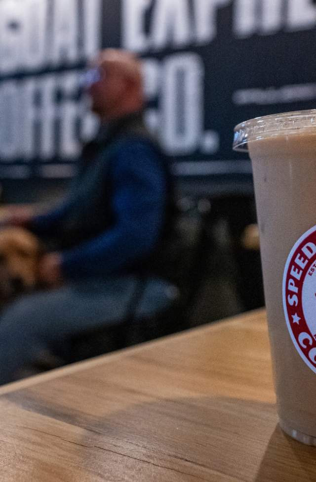 A clear plastic cup with iced coffee sits on a wooden table in a cafe. The logo reads "Speed Goat Espresso." A blurred background shows a person and a dog.