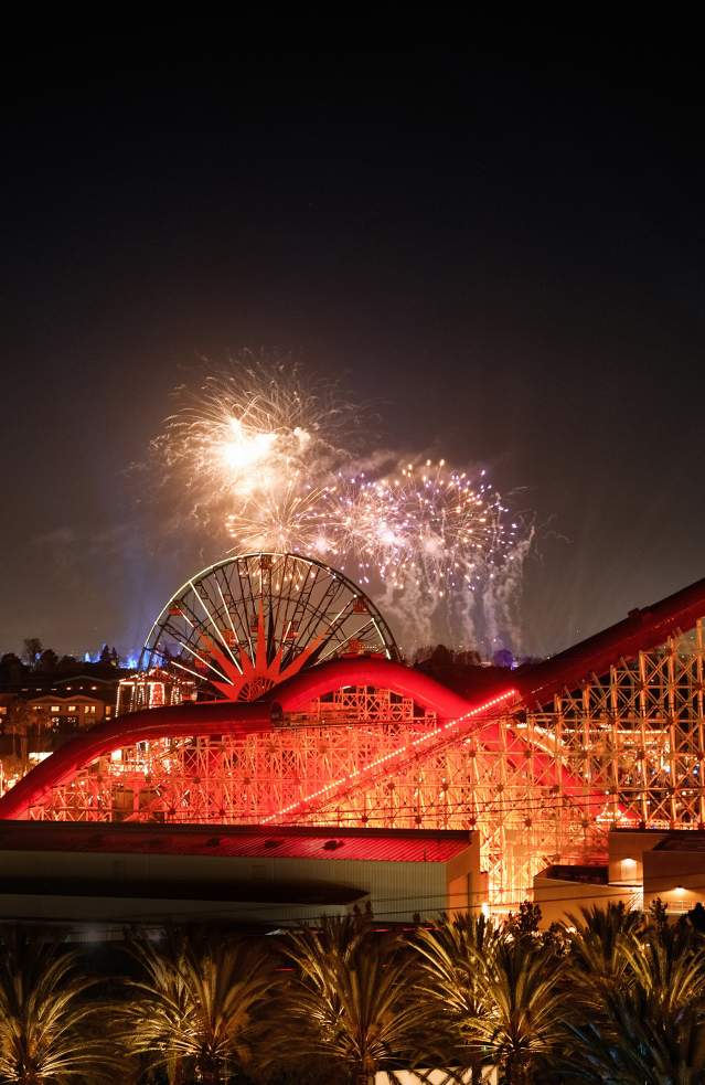 Disney fireworks lighting up the night sky over a brightly illuminated roller coaster and Ferris wheel at a theme park in Anaheim, California.