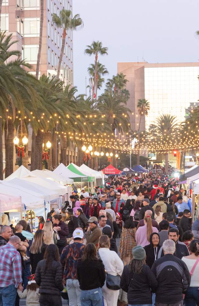 Downtown Anaheim shopping district featuring a bustling outdoor market with vendor tents, shoppers walking along a palm‑lined street, festive string lights overhead, and local businesses surrounding the lively event atmosphere.