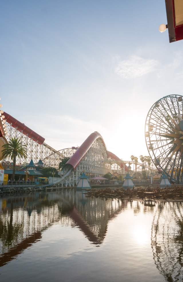 Sunrise at Disney California Adventure Park with a large Ferris wheel and roller coaster, reflected in a serene lake surrounded by palm trees.