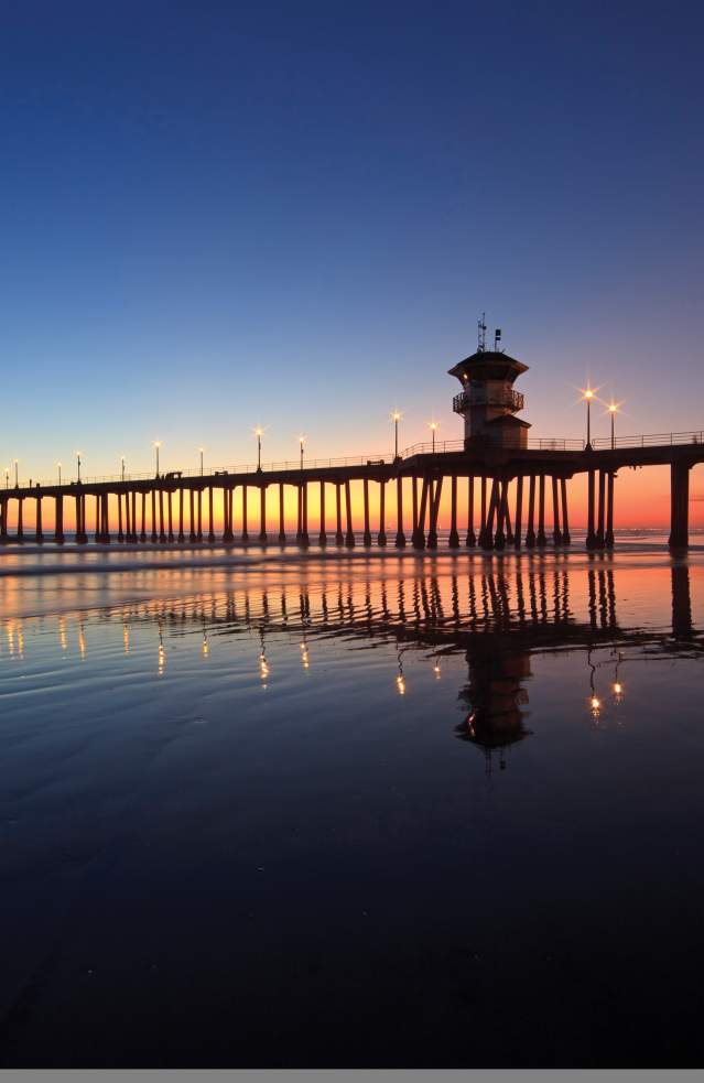 Huntington Beach pier at sunset with lights reflecting on wet sand, extending into calm ocean waters under a gradient sky of deep blue and warm orange hues.