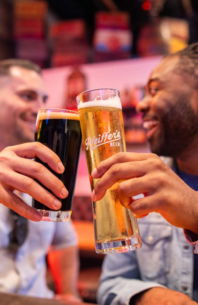 Pfeiffer Brewing Company in Anaheim, California, featuring two patrons raising glasses of craft beer in a toast inside a warm, lively brewery setting with wood accents and ambient lighting.