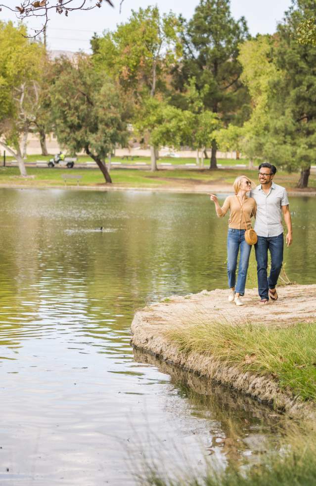 Yorba Regional Park in Anaheim featuring two people walking along a grassy shoreline beside a calm lake, with trees, open green space, and gentle ripples on the water under a bright sky.