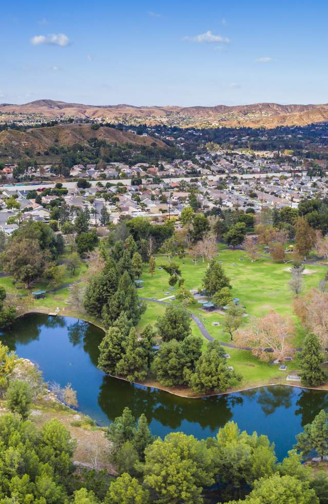 Yorba Regional Park in Anaheim featuring a wide aerial view of scenic lakes, tree-lined green spaces, walking paths, and nearby neighborhoods with rolling hills in the distance under a clear blue sky.