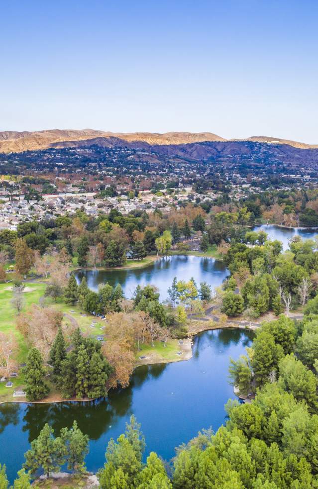 Yorba Regional Park in Anaheim featuring an aerial view of tree-lined lakes, winding walking paths, open green spaces, and surrounding residential neighborhoods under a clear blue sky.