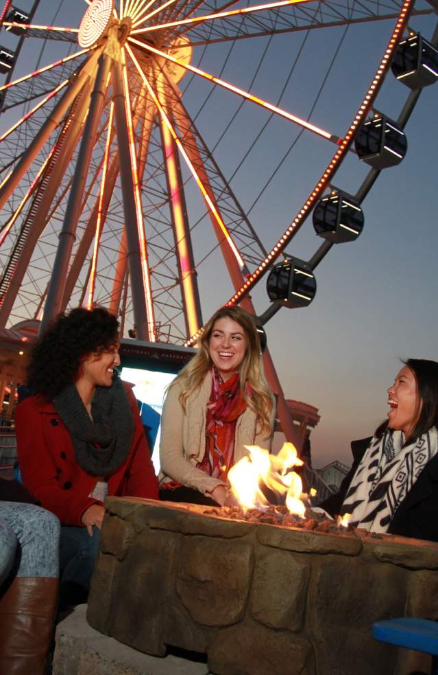 Group seated in red Adirondack chairs around a stone fire pit beside the waterfront in Prince George’s County, Maryland, with a brightly lit Ferris wheel rising in the background at dusk.