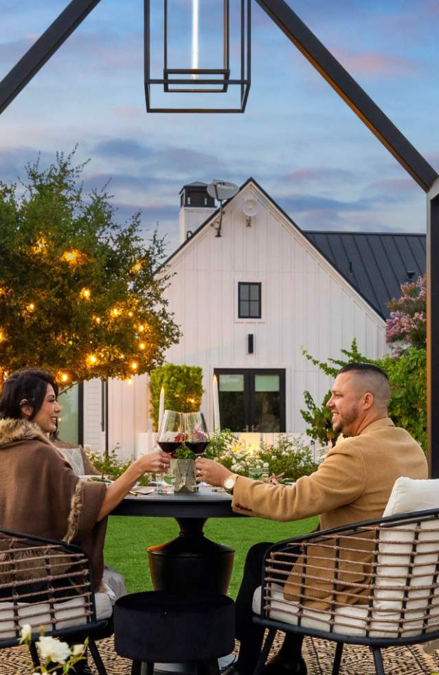 Man and woman sitting sipping wine at small table at sunset with beautiful white inn in background