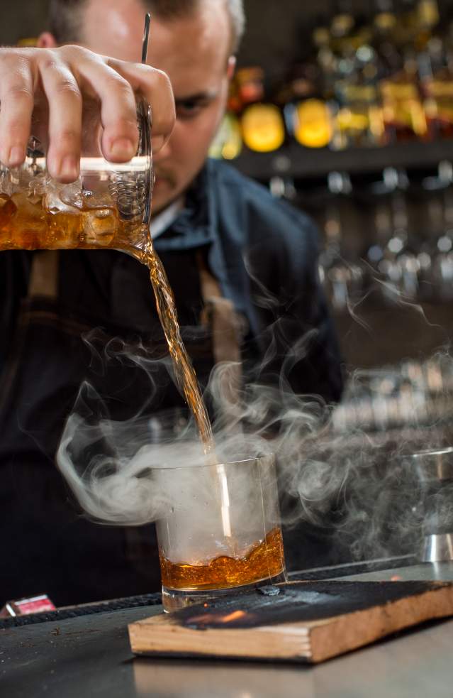 Bartender pouring a smoking Cedar cocktail at a bar with aromatic presentation