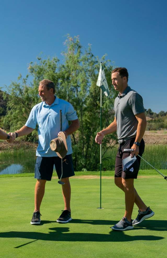 Three men in golf attire shaking hands on a sunny golf course, surrounded by lush greenery and a clear blue sky.