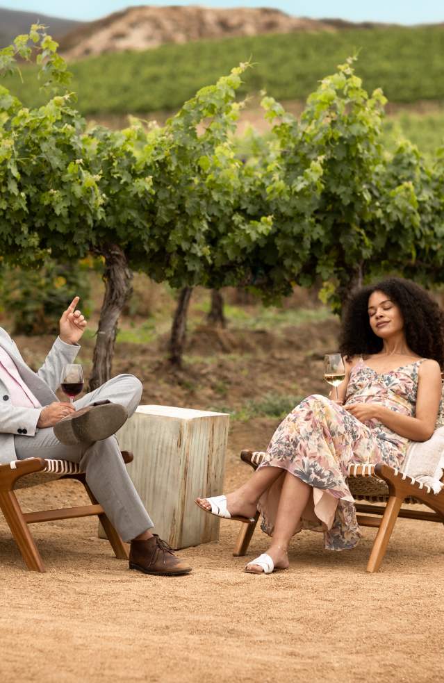 Man and woman sitting in chairs with rolling vineyards behind them, relaxing with eyes closed.
