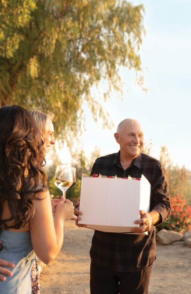 Several people stand outside a vineyard conversing, enjoying a glass of wine