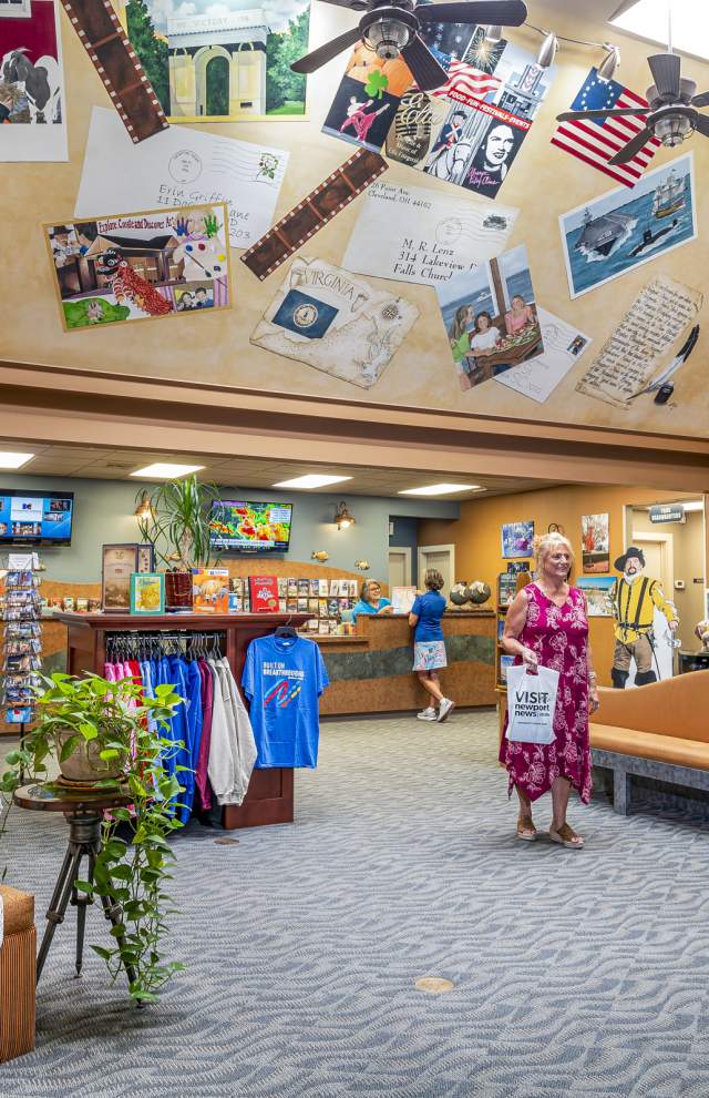 A woman is sitting reading the visitor guide magazine while another woman shops in a vaulted room with an artistic ceiling covered in places around Newport News