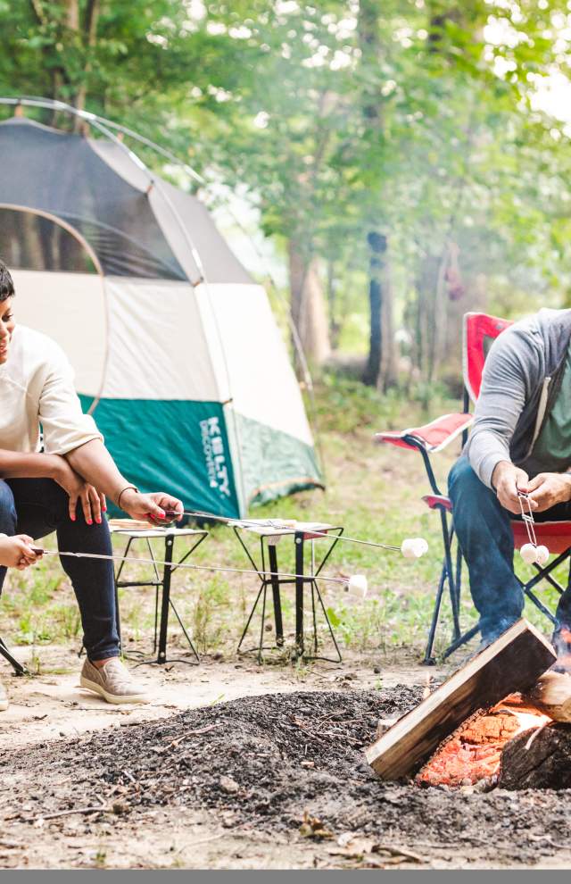 Family of 4 roasts marshmallows over a fire in front of a tent in the woods