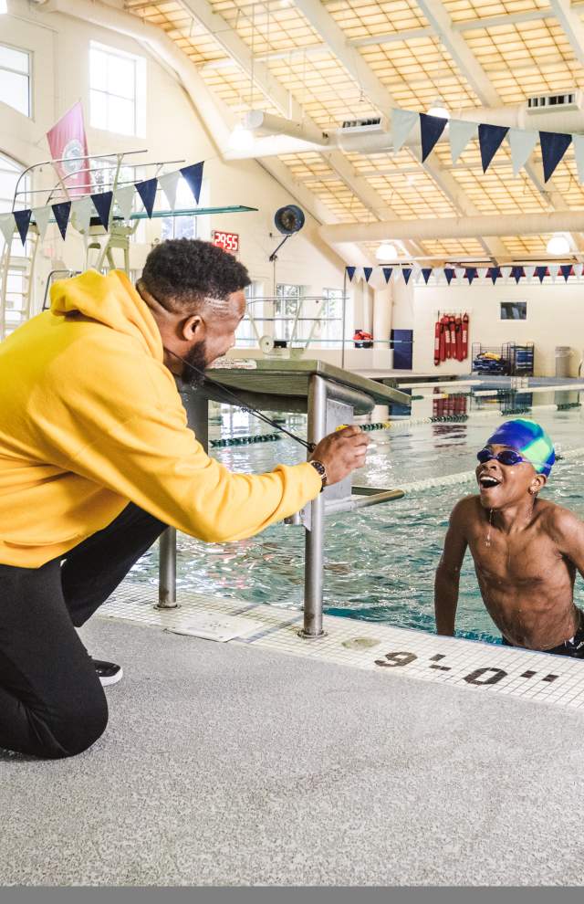 child emerging from a large pool with google and swim cap while encouraging adult shows the child their time