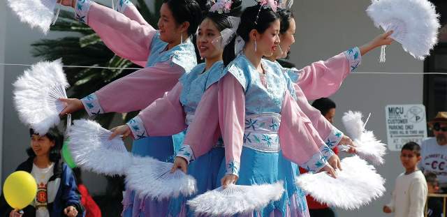 4 women of Asian descent dressed in native costumes in light blue and pink, holding white feather fans