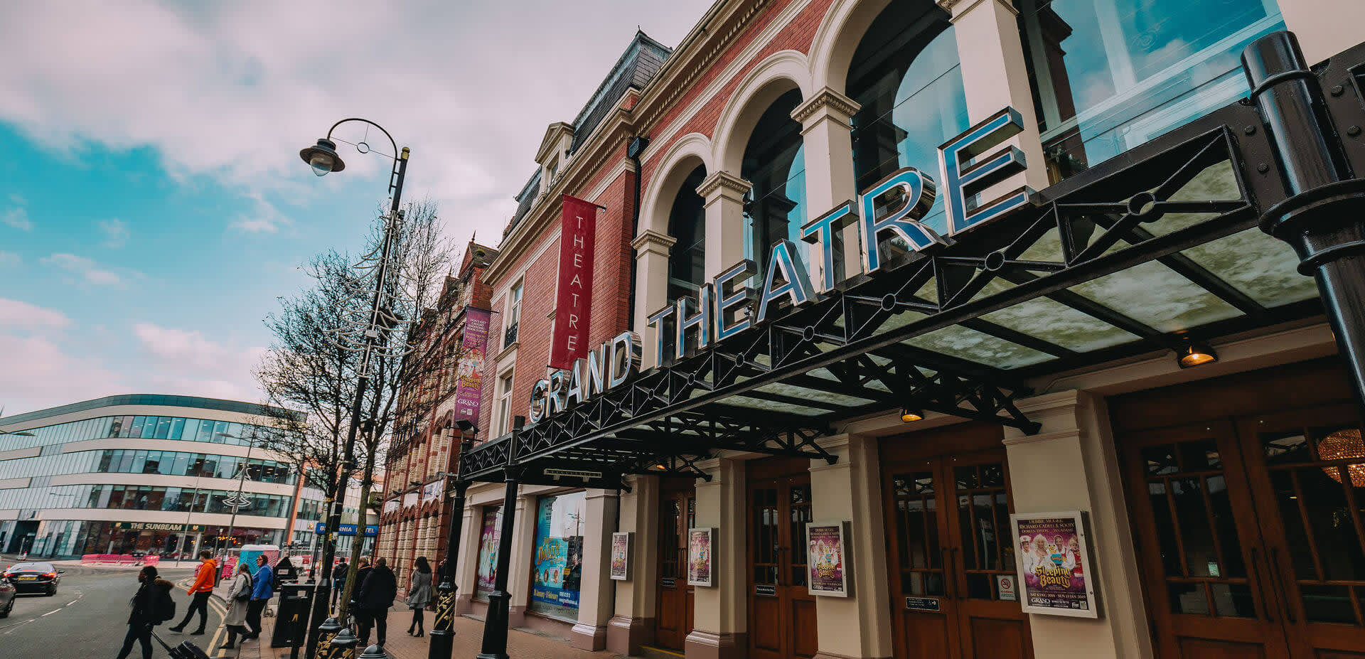 Exterior of the Grand Theatre in Wolverhampton. Large letters GRAND THEATRE on top of a metal canopy covering the 4 double door entrance to the theatre on a busy street.