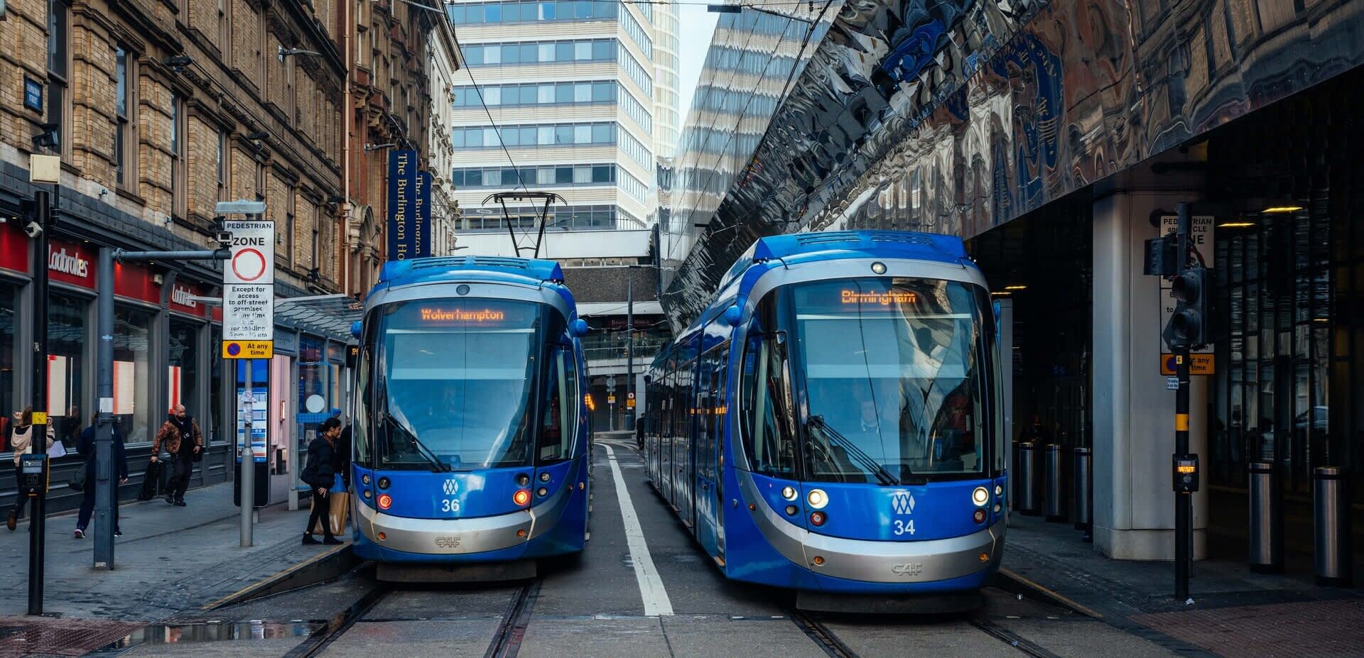 Two blue trams, parked side by side in a street, let passengers embark.