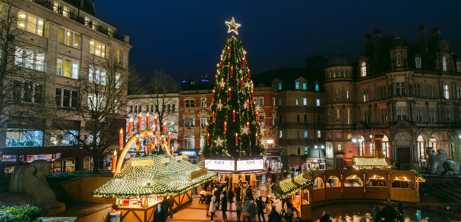 A tall Christmas tree stands in a square in Birmingham at night, the lights are lit and people are gathered round its base at Christmas market stalls