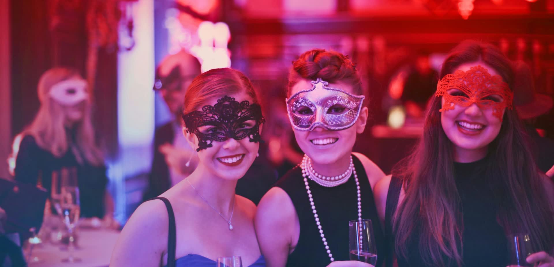 Three people wearing masquerade masks smile at the camera in a nightclub