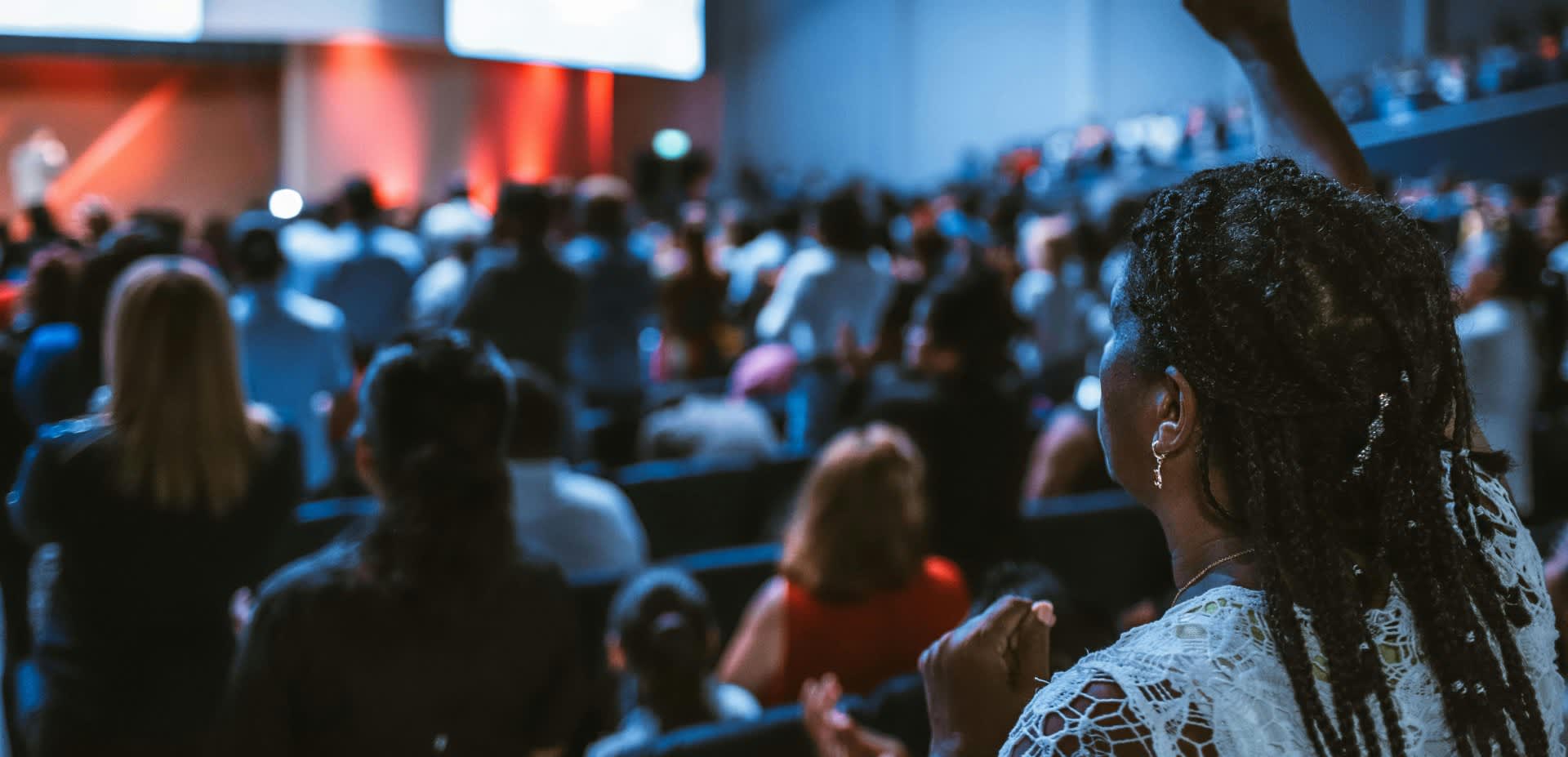 A woman raises her hand in a room full of people with a stage at he front