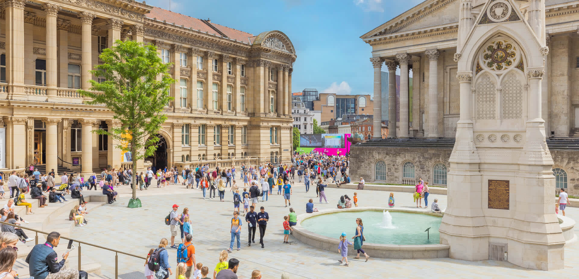 Groups of people sitting outside in summer clothes. Surrounded by Chamberlain Square fountain and Birmingham Museum and Art Gallery