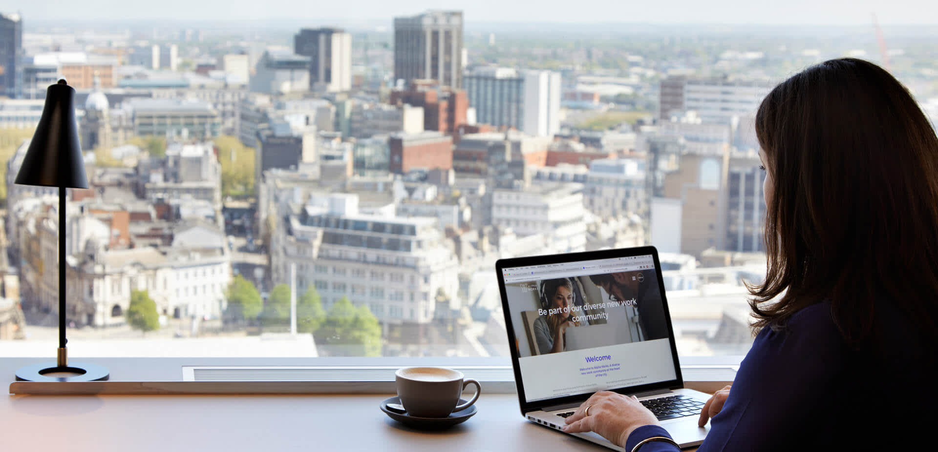 Girl on laptop with a hot drink and view of Birmingham's skyline out of the window