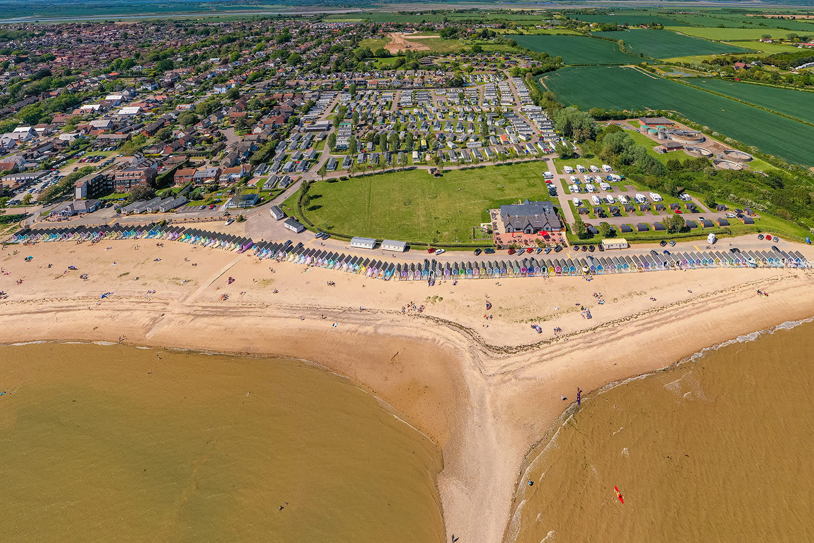 Mersea's famous pastel beach huts are captured from a distance in a drone shot that highlights the nearby sandy beaches and green fields